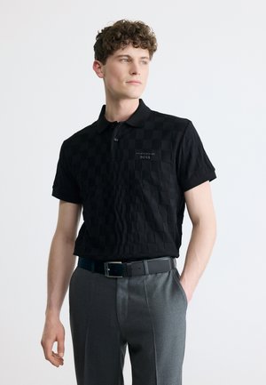 Tânăr purtând un polo Porsche BOSS negru în carouri și pantaloni gri, stând cu o mână în buzunar și privirea îndreptată lateral.