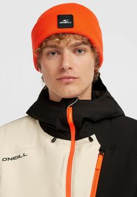 Oranje gebreide beanie met zwart logo-patch, gedragen met een multi-kleurige jas met zwart, beige en een oranje ritssluiting detail.