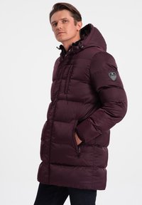 Giubbotto imbottito bordeaux con cappuccio, dotato di chiusura a zip, tasche frontali e un toppe con logo sul braccio superiore. Tessuto liscio e isolato.