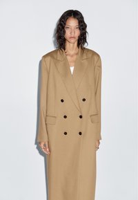 Double-breasted coat in light brown fabric, featuring large black buttons, notched lapels, and side pockets. Long, straight silhouette.