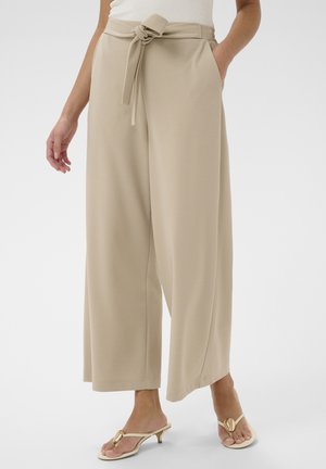 Pantalon beige à jambes larges avec ceinture en tissu nouée à la taille, porté avec un haut blanc et des sandales à talons ouvertes beiges, modèle debout avec une main dans la poche.