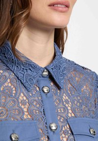 Chemise en dentelle bleu clair avec un motif floral détaillé, col à boutons et boutons argentés sur le devant et les poches poitrine.