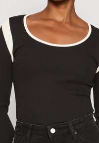 Schwarzes Langarmoberteil mit Rundhalsausschnitt und geripptem Design, mit cremefarbenen Akzenten an den Schulternähten. Passt gut zu schwarzen Jeans.