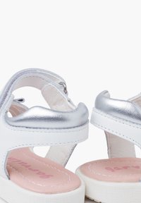Białe sandały z metalicznymi srebrnymi akcentami, odkryty tył i wyściełana różowa wkładka z białymi detalami przeszycia.