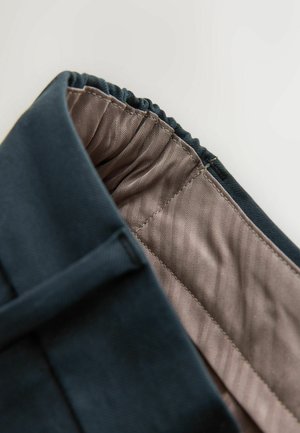 Tumedates sinises riidis on kokkutõmbunud vöökoht, mis paljastab heledat pruuni sisemust, millel on herringbone muster ja sile tekstuur.