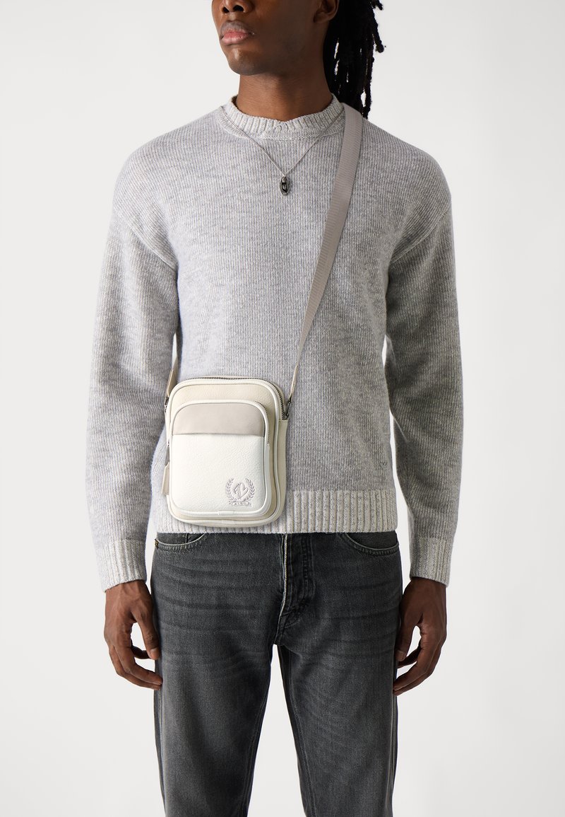 Borsa a tracolla in pelle bianca e grigia con chiusura a zip, tasca frontale e dettaglio del logo; abbinata a un maglione a coste grigio chiaro.