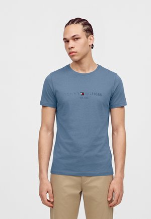 Млад мъж с вплетена коса, облечен в синя тениска с къс ръкав Tommy Hilfiger и бежови панталони, стоящ срещу едноцветен фон.