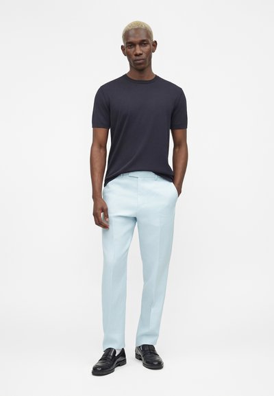 Άνδρας με κοντά ξανθά μαλλιά που φοράει μπλε σκούρο μπλουζάκι, ανοιχτό μπλε παντελόνι και μαύρα επίσημα παπούτσια, στέκεται με το ένα χέρι στην τσέπη.