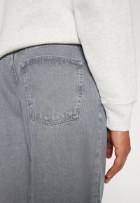 Gray denim jeans displayed from the side, featuring a back pocket and a light gray sweatshirt cuff visible above. Textured fabric and simple design.