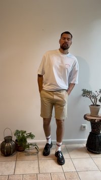 T-shirt bianca con testo stampato rosso, pantaloni corti beige, scarpe nere con suole spesse e calzini bianchi con strisce rosse e nere. Ambientazione interna.
