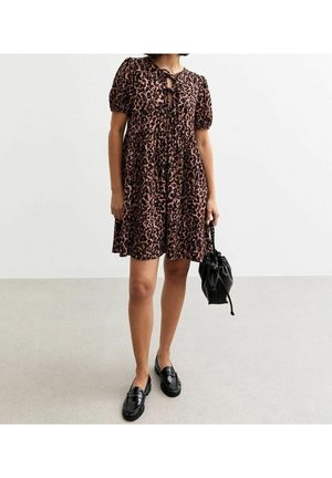 Kobieta w krótkiej sukience w panterkę z wiązaniami z przodu, trzymająca czarną torebkę na troczki i nosząca czarne mokasyny na jasnym tle.