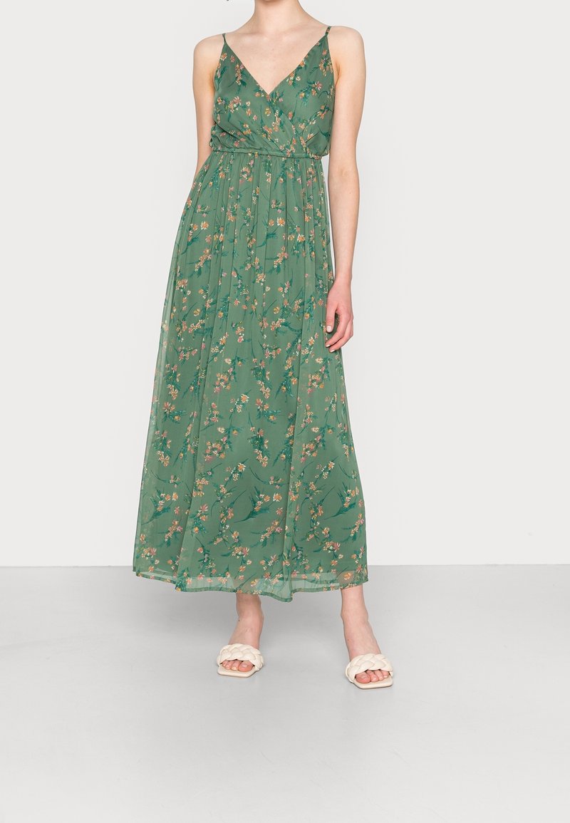 Femme portant une robe maxi verte à fleurs sans manches avec bretelles spaghetti et sandales slides blanches tressées.