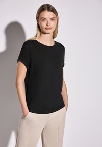 Sort t-shirt med korte ærmer, der har et tekstureret strikket overpanel; glat stof nedenunder, parret med beige bukser.