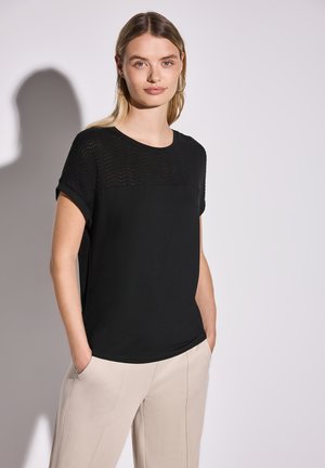 Sort t-shirt med korte ærmer, der har et tekstureret strikket overpanel; glat stof nedenunder, parret med beige bukser.