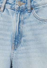 Blugi din denim albastru deschis, în prim-plan, care arată nasture metalic, fermoar frontal cu cusături și o parte din buzunarul din față.