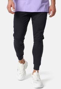 Persona vistiendo pantalones deportivos negros y zapatillas blancas, caminando con un pie ligeramente levantado, con una camiseta de color púrpura claro.