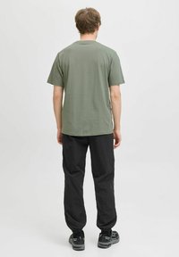 Camiseta de algodón verde con mangas cortas, combinada con pantalones negros de cintura elástica y zapatillas oscuras, vista desde la parte posterior.