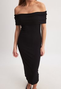 Melnā ribota kleita, kas tiek valkāta bez pleciem, ar pieguļošu siluetu, līdz ceļiem garu dizainu un gludu tekstūru. Iezīmē taisnu kakla izgriezumu un vidēja garuma piedurknes.