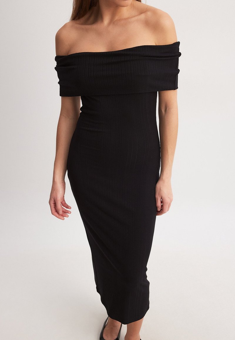 Melnā ribota kleita, kas tiek valkāta bez pleciem, ar pieguļošu siluetu, līdz ceļiem garu dizainu un gludu tekstūru. Iezīmē taisnu kakla izgriezumu un vidēja garuma piedurknes.