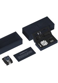 Die marineblaue Verpackung enthält eine silberne Uhr mit blauem Ziffernblatt, Gummiband und zusätzlichem Werkzeug, das in einem konturierten schwarzen Einsatz präsentiert wird.