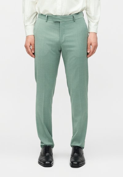 Άνδρας που φοράει ανοιχτοπράσινο παντελόνι ραμμένο στα μέτρα του, λευκό πουκάμισο μέσα, και μαύρα δερμάτινα παπούτσια, στέκεται μπροστά σε μονόχρωμο φόντο.