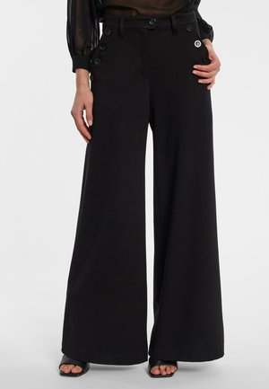 Black wide-leg pants with high waist and side buttons worn with open-toe black heeled sandals and sheer long-sleeve top.