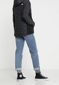 Parka nera imbottita con un motivo esagonale testurizzato, abbinata a jeans azzurri con risvolto e sneakers nere alte con dettagli bianchi.