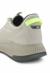 Helle graue Sneaker aus strukturiertem Material, ausgestattet mit einer kontrastierenden neongrünen Fersenschlaufe, erhöhten Sohle mit Rillenoptik und Logo-Branding.