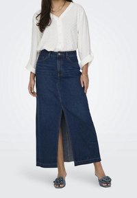 Długa spódnica dżinsowa z rozcięciem z przodu, w ciemnym kolorze, wysoka talia i kieszeniami, zestawiona z białą bluzką z guziczkami w kropki.