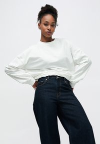 Λευκό πουκάμισο με μακριά μανίκια συνδυασμένο με σκούρο μπλε ψηλόμεσο τζιν, με ίσια μπατζάκια και κλασικό σχέδιο με πέντε τσέπες.