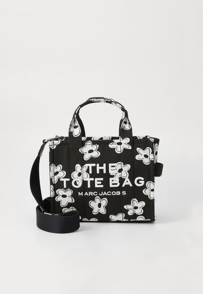 Черна чанта тип торба с бял флорален принт, къси дръжки, свалящ се презрамка и текст "THE TOTE BAG MARC JACOBS" на предната част.