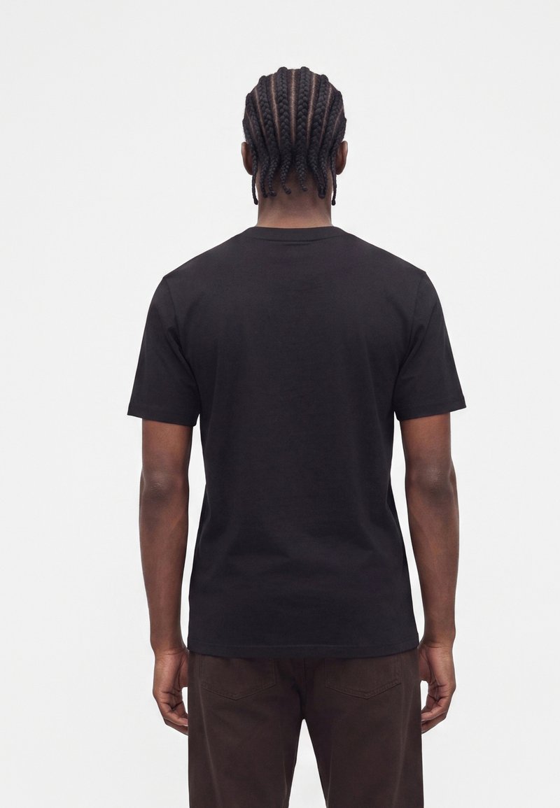 Mann mit geflochtenem Haar, der ein schlichtes schwarzes T-Shirt mit kurzen Ärmeln und dunkle Hose trägt, steht mit dem Rücken zu einem weißen Hintergrund.