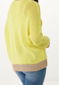 Yellow knitted sweater with a loose fit, featuring a beige ribbed hem. Soft texture, drop shoulders, and a wide neck. Worn with blue jeans.