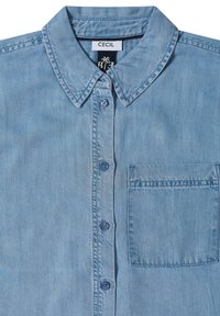 Chemise en jean bleu clair à boutons, avec col et poche poitrine, étiquetée "CECIL" à l'intérieur de l'encolure.