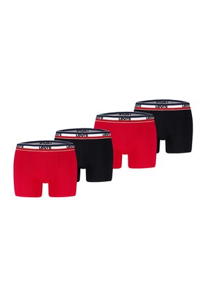 Muskolöser männlicher Oberkörper, der rote Levi's Boxershorts mit weißem und marineblauem Bund trägt, steht vor einem schlichten hellen Hintergrund.