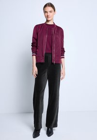 Burgundrote Bomberjacke mit gerippten Bündchen, kombiniert mit weiten schwarzen Cordhosen. Das Model trägt schwarze spitze Stiefel. Einfacher Hintergrund.