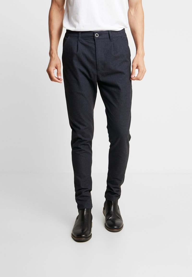 Homme portant un pantalon slim foncé et des bottines en cuir noir, debout devant un fond clair uni.