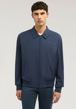 Junger Mann trägt eine marineblaue Jacke mit Reißverschluss, Kragen und gerippten Bündchen, steht vor einem einfachen hellen Hintergrund.