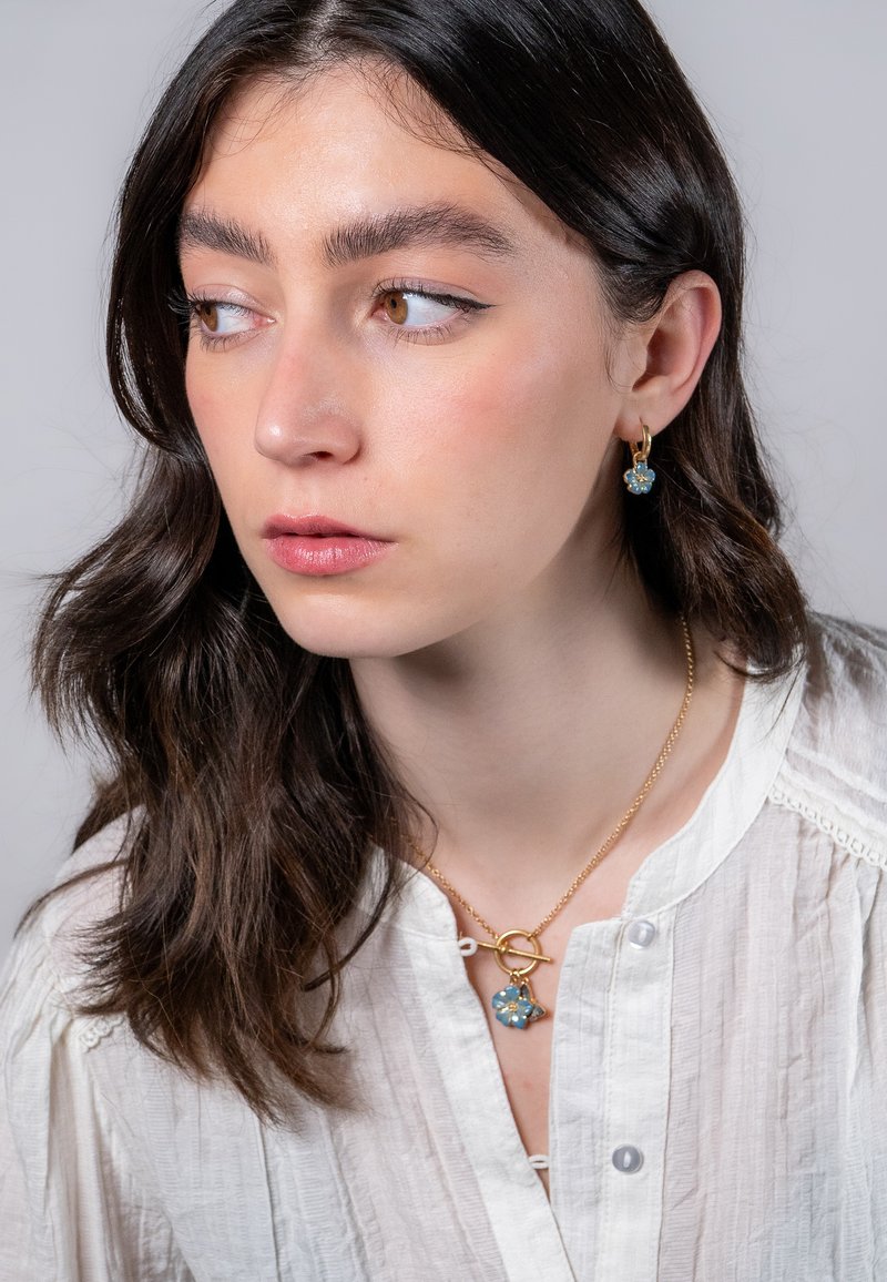Collana in oro con pendente a forma di fiore blu. Modella che indossa orecchini a cerchio in oro con accenti blu simili. Blusa bianca con texture.