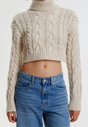 Lille cremet kabelstrikket sweater med høj krave, der har en tykkere tekstureret garn. Stylet med højtaljede blå denimjeans.