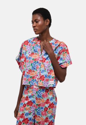 Femme aux cheveux courts portant un ensemble assorti coloré à motifs floraux, composé d'un haut à manches courtes et d'un pantalon à coupe décontractée, debout devant un fond uni.