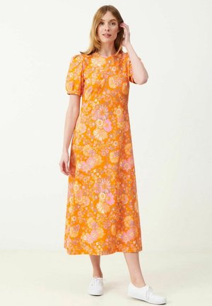 Nainen, joka pukeutuu oranssinväriseen kukalliseen midi-mekkoon, jossa on lyhyet pörröiset hihat, ja valkoiset tennarit, seisoo yksinkertaista vaaleaa taustaa vasten.