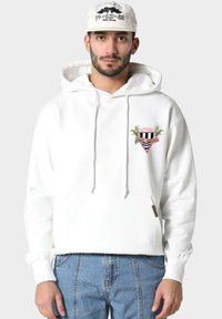 Uomo con felpa bianca con grafiche di una palma e di una macchina da corsa, jeans blu e berretto bianco con la scritta "1988" e un design di un pipistrello.