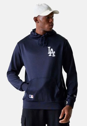 Mann trägt einen marineblauen LA Dodgers Hoodie und eine weiße Kappe, steht mit einer Hand in der Tasche und blickt nach rechts.