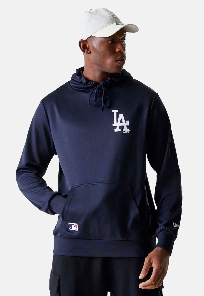 Homme portant un sweat à capuche bleu marine des LA Dodgers et une casquette blanche, debout avec une main dans la poche, regardant vers sa droite.