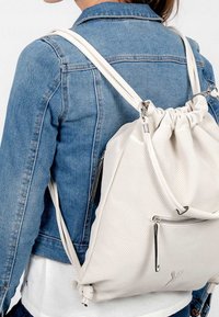 Sac à dos perforé blanc avec un haut resserré, des bretelles réglables et une poche zippée à l'avant. Texture lisse et accents en métal argenté.