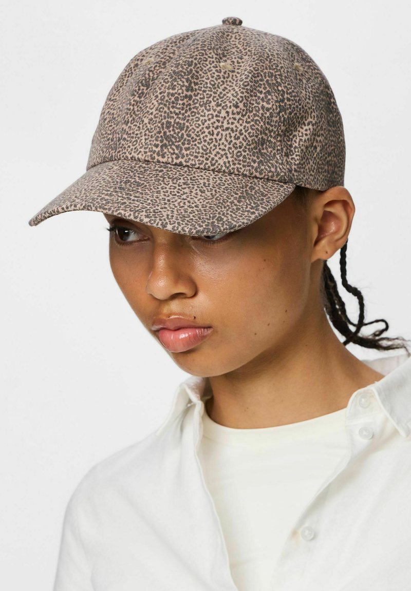 Personne aux cheveux tressés portant une casquette de baseball claire à motif animalier et une chemise blanche boutonnée, regardant légèrement vers le bas et sur le côté.