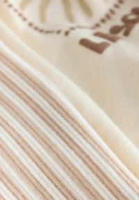 Krema in svetlo bež tkanina z vodoravnimi črtami, z rjavimi potiski. Mehka tekstura poudarja vizualno globino in vzorec.