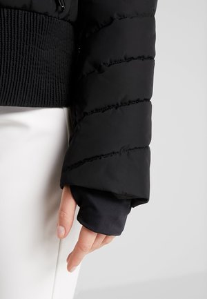 Hand trägt schwarzen Steppjackenärmel mit geripptem Bund über schwarzem Innensleeve, ruht neben weißer Hose auf hellem Hintergrund.