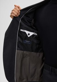 Zwarte blazer met een gestructureerde buitenkant, voorzien van een zachte binnenvoering met een herhalend logopatroon en een verborgen zak met een witte klep.